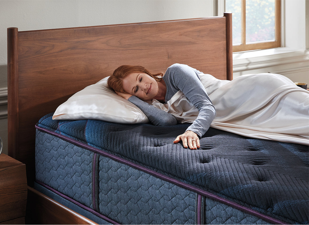 Woman laying on her right side with her head on a pillow as she rubs the top of a Beautyrest World Class mattress. 